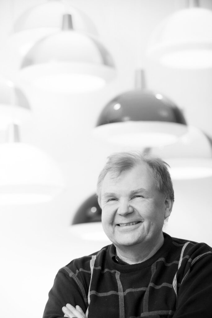 En brett leende äldre man med ljust hår och svart tröja med gråa ränder i rutmönster. Bakom och ovanför honom hänger flera taklampor med en modern design.
