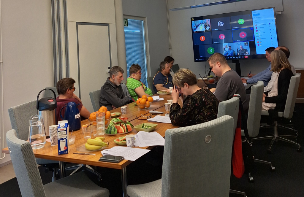 10 personer samlade kring ett mötesbord. På väggen en stor skärm med kontakt till distrikten online.
